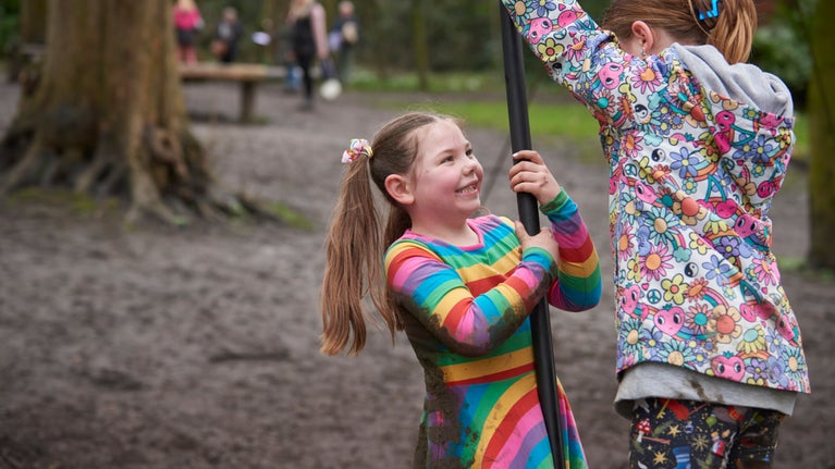 Two girls on the cantilever swing in the woodland play area at Nostell. One girl wears a rainbow coloured dress covered in mud.